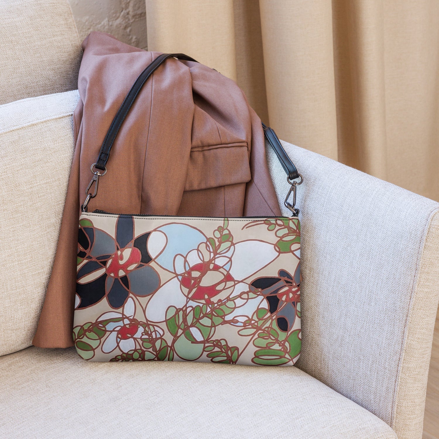 A purse with a black strap, in the corner of a couch leaning against a leather jacket. The purse has an abstract flower design in burnt orange, black and white 